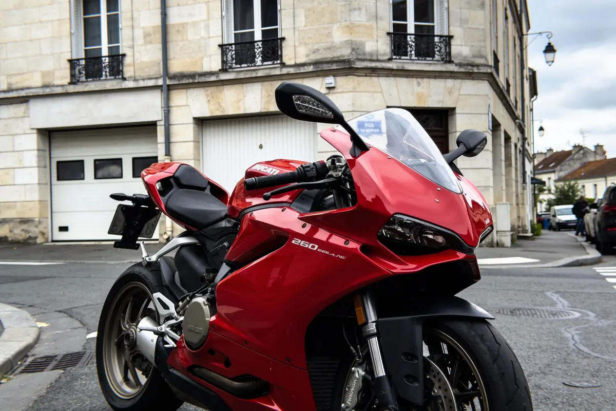 Moto rouge garée dans une rue, devant un immeuble en pierre, journée nuageuse.