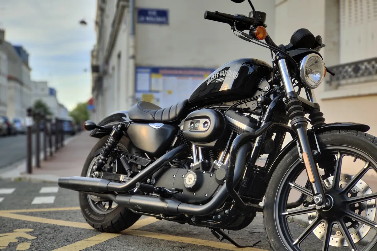 Moto Harley-Davidson noire garée sur trottoir urbain, bâtiments arrière-plan flou, ciel partiellement couvert.