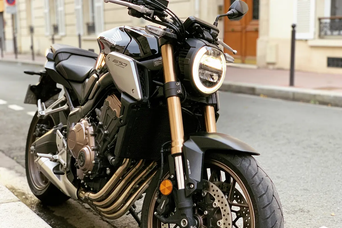 Moto noire stationnée dans une rue, design moderne avec fourche dorée et phares ronds allumés.