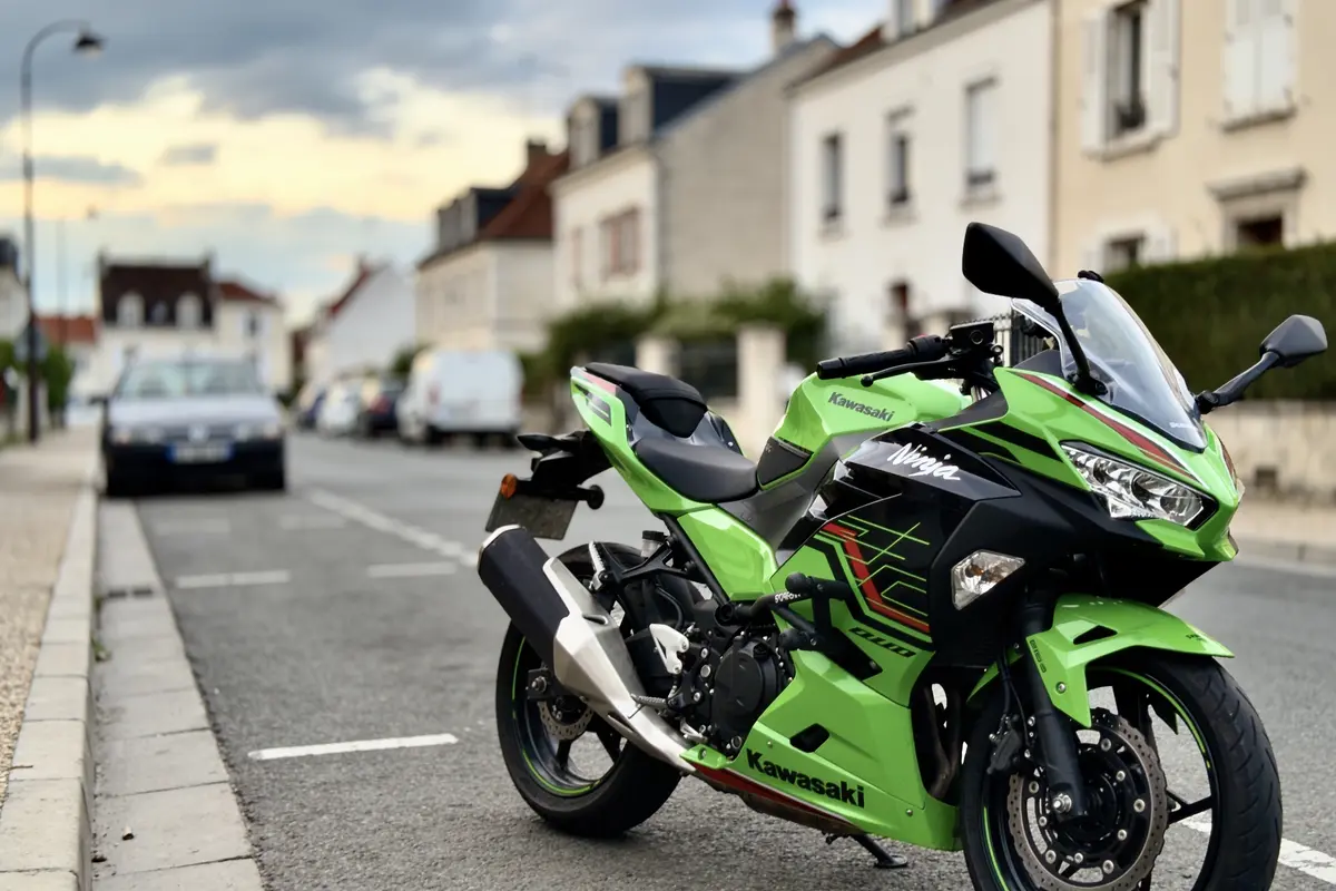 Moto Kawasaki verte garée sur une rue, maisons et voitures en arrière-plan sous ciel nuageux.