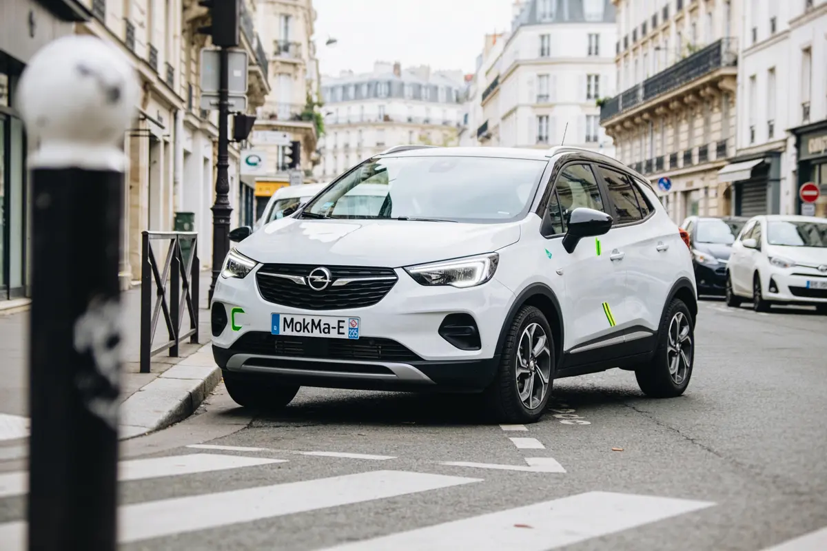 Voiture blanche, Opel, garée en ville, jour, bâtiments classiques, rue calme, plaque "MokMa-E".