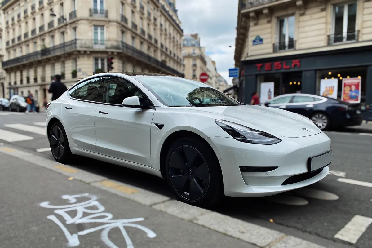 Une voiture blanche garée en ville devant un magasin Tesla, bâtiments en arrière-plan.