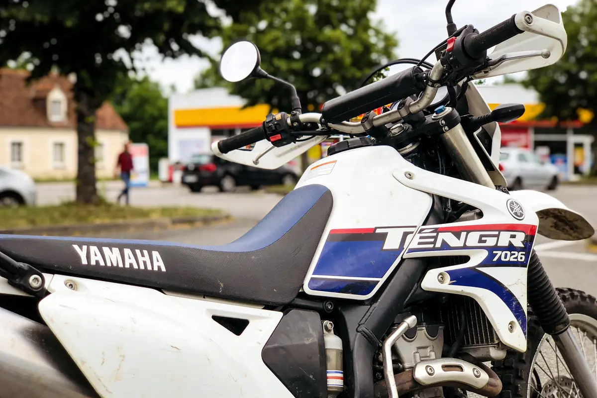 Moto Yamaha Ténéré blanche et bleue stationnée devant station-service, arbres et bâtiments en arrière-plan.