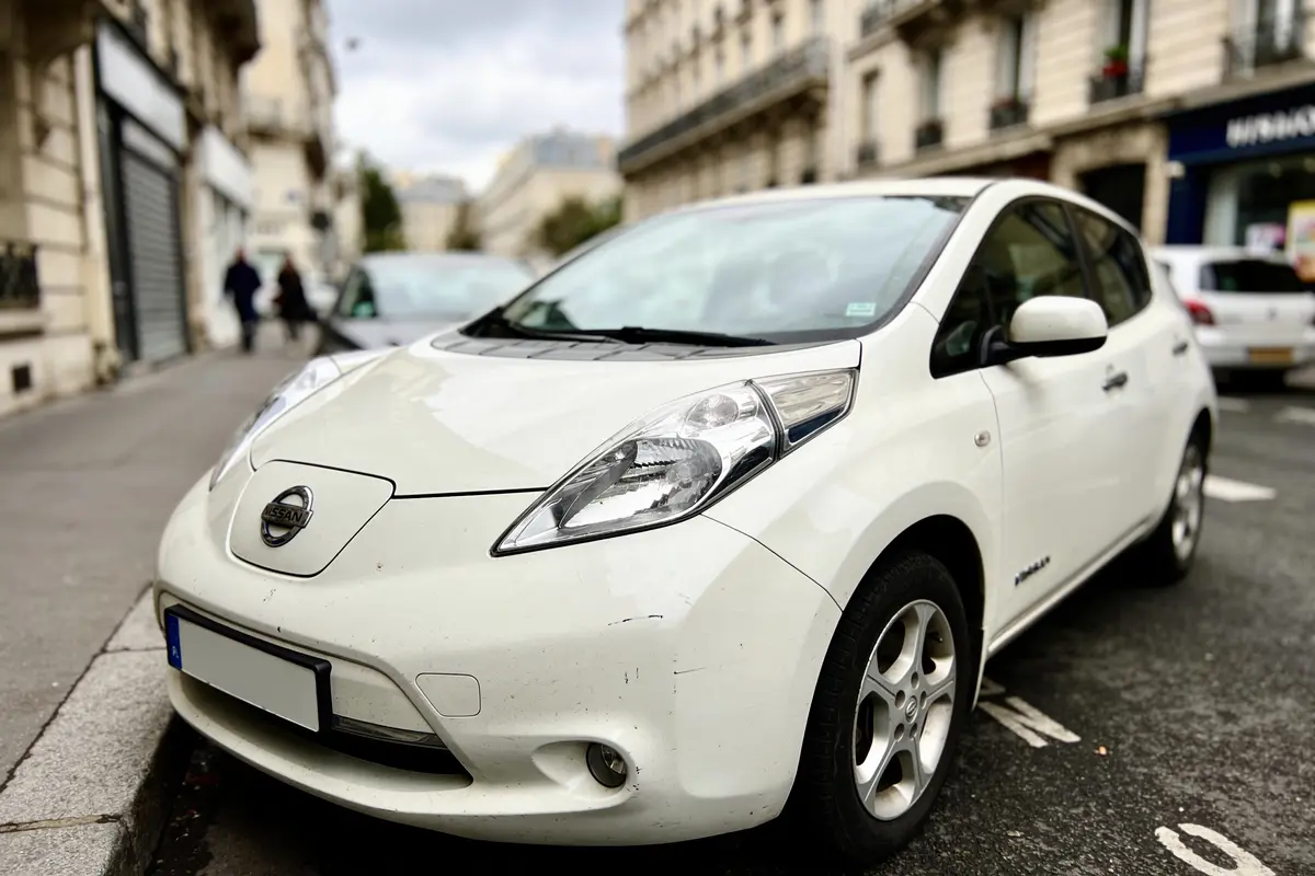 Voiture blanche garée dans une rue urbaine, bâtiments en arrière-plan flou.