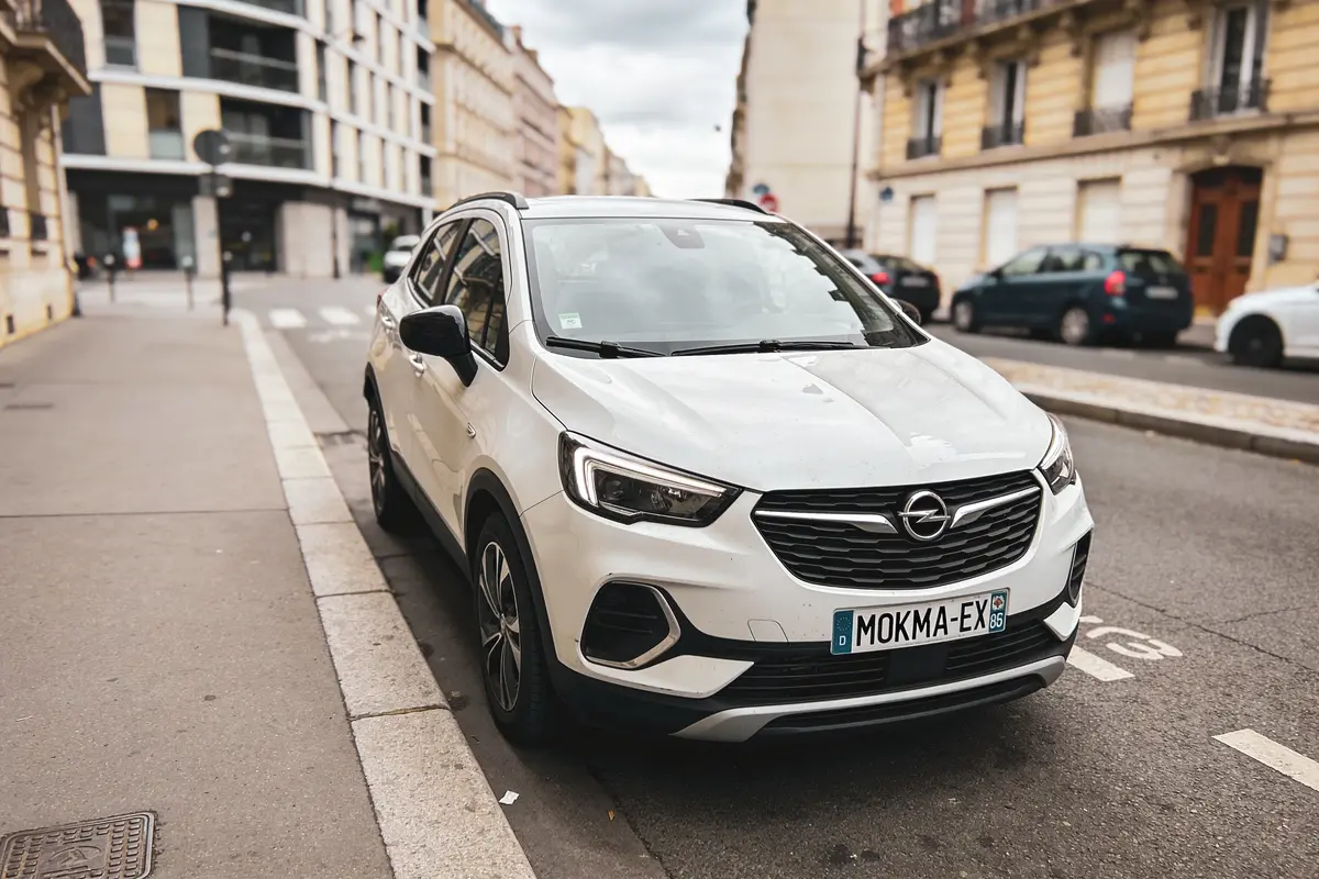 Voiture blanche Opel garée en bord de trottoir, immeubles en arrière-plan urbain.