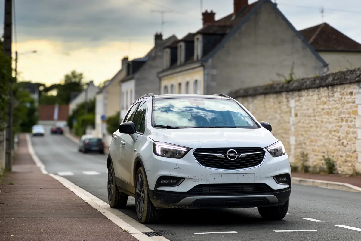 SUV blanc Opel sur une route de campagne pavée, maisons en arrière-plan, ciel nuageux.