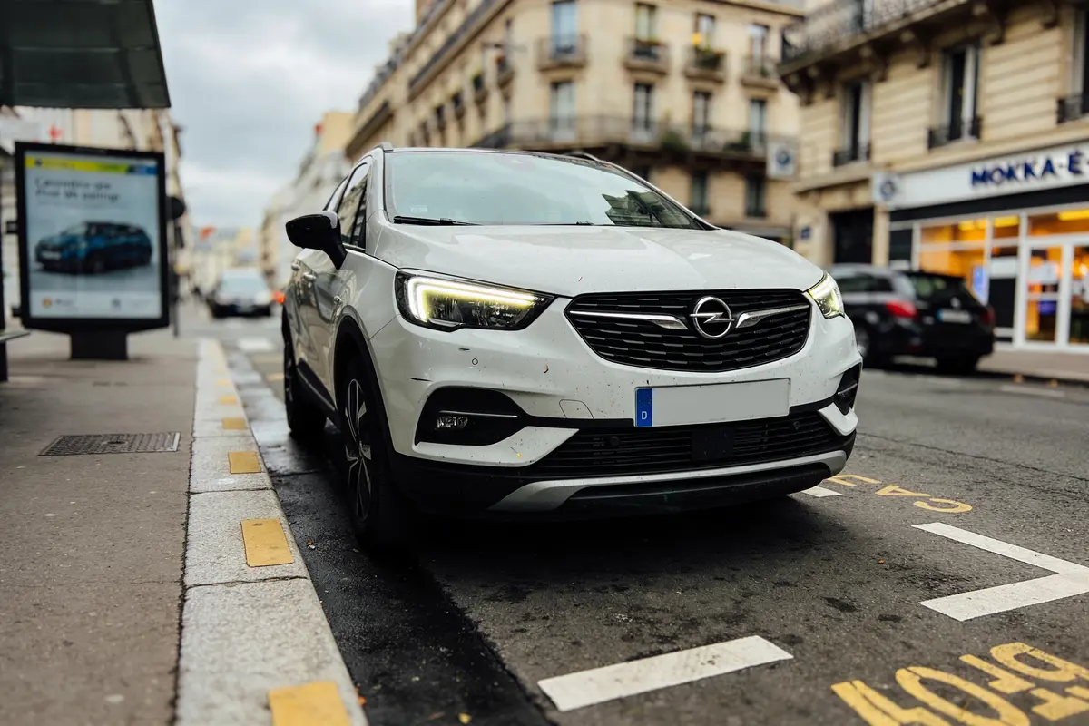 Voiture blanche Opel garée sur une rue urbaine, bâtiments en arrière-plan, panneau publicitaire visible.