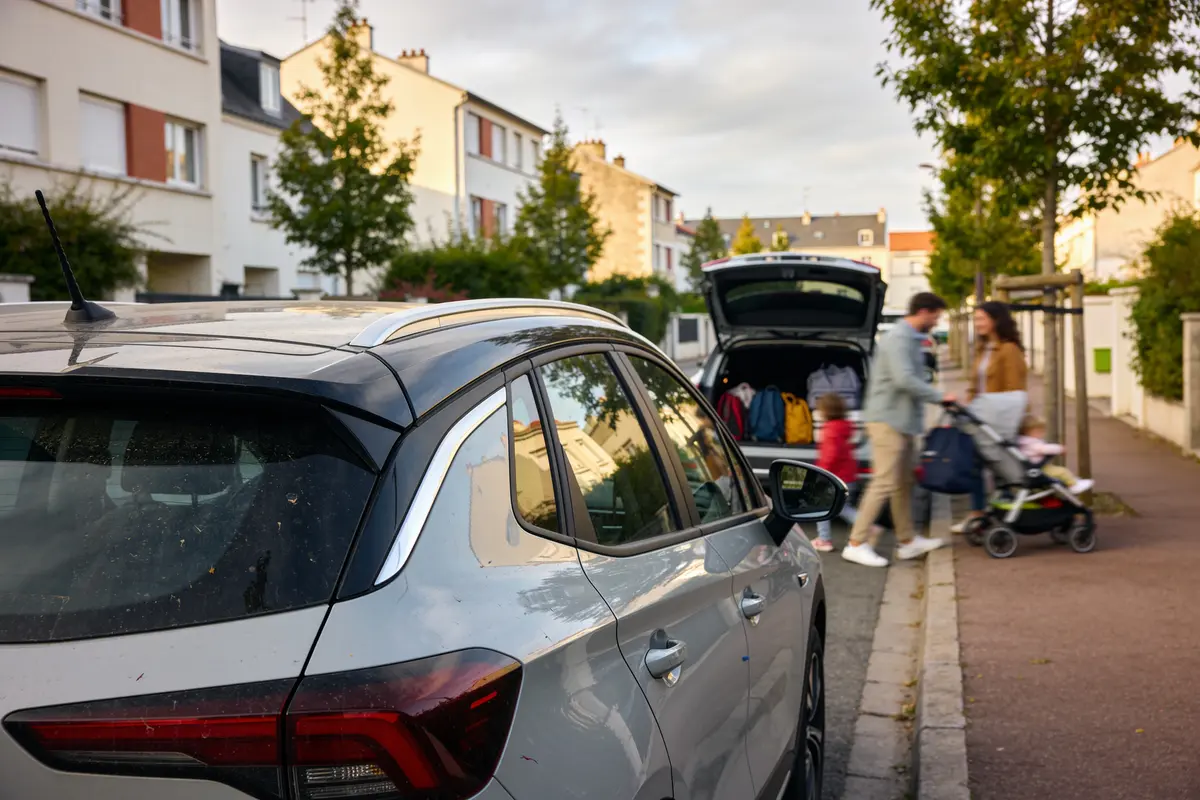 Voiture garée dans rue résidentielle, famille se préparant au départ, coffre ouvert.