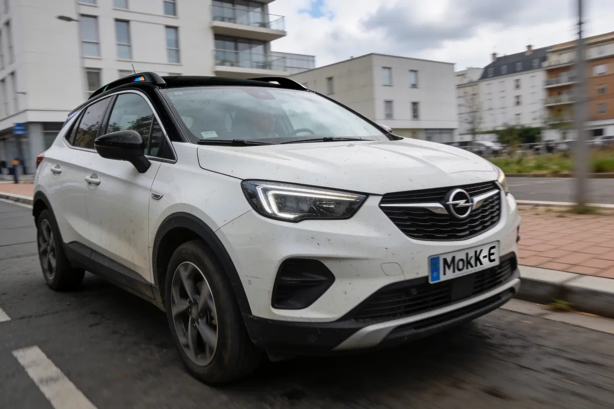 Voiture blanche Opel en mouvement, sur route urbaine, bâtiments modernes en arrière-plan. Plaque "MokK-E".