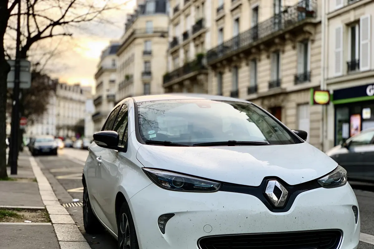 Voiture blanche Renault stationnée dans une rue urbaine, bâtiments résidentiels en arrière-plan par temps nuageux.